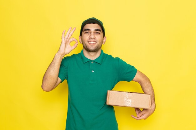 Front view young male courier in green shirt green cap smiling and holding delivery box showing alright sign on the yellow background delivering service color