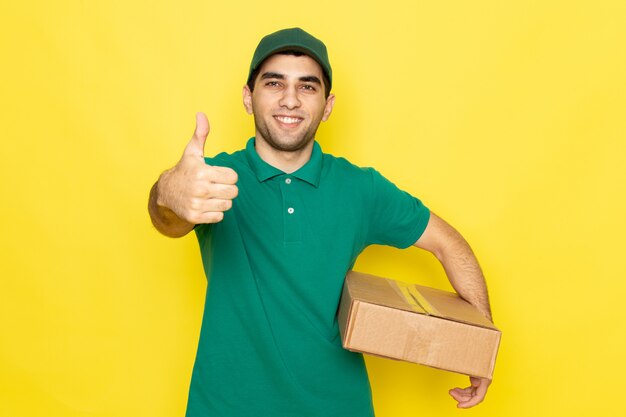 Вид спереди молодой мужчина-курьер в зеленой рубашке с зеленой кепкой улыбается и держит коробку доставки на желтом фоне.