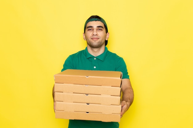 Вид спереди молодой мужчина-курьер в зеленой рубашке с зеленой кепкой улыбается и держит коробки доставки на желтом фоне, предоставляя цвет услуг