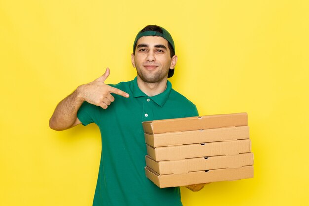 笑顔の緑のシャツの緑の帽子の正面の若い男性宅配便とサービスの色を提供する黄色の背景の仕事の配達箱を保持