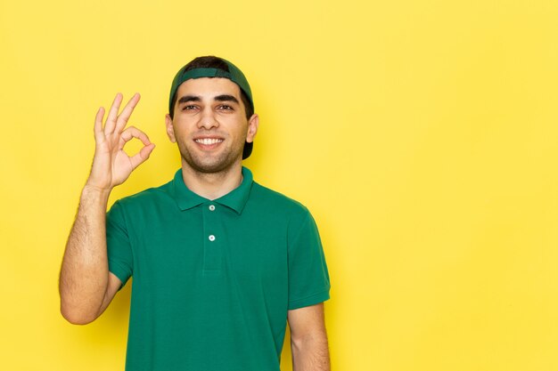 Front view young male courier in green shirt green cap smiling and showing alright sign smiling on the yellow background job delivering service color