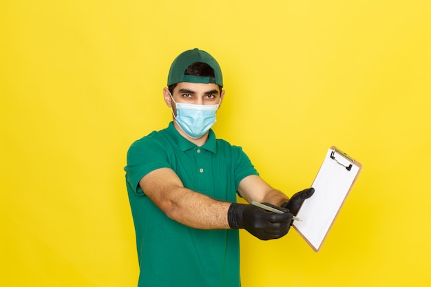 Front view young male courier in green shirt green cap writing down notes on the yellow background delivering service color job work