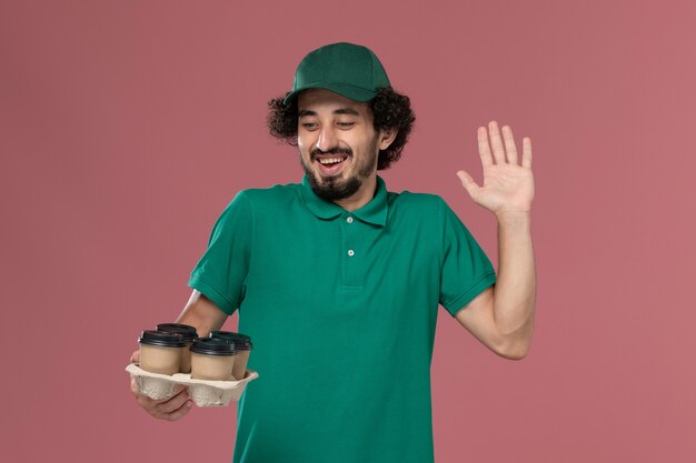 緑の制服と薄ピンクの背景サービス制服配達労働者に茶色の配達コーヒーカップを保持している岬の正面図若い男性宅配便