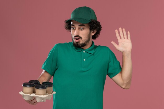 Front view young male courier in green uniform and cape holding brown delivery coffee cups on the light-pink background service uniform delivery worker