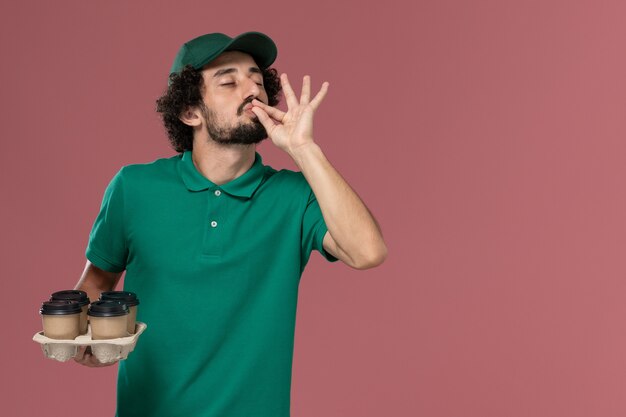 緑の制服とピンクの背景に茶色の配達コーヒーカップを保持している岬の正面図若い男性の宅配便制服配達仕事の仕事