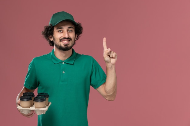 Вид спереди молодой курьер-мужчина в зеленой форме и плаще, держащий коричневые кофейные чашки доставки на розовом столе, работник службы доставки униформы