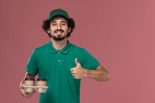 Front view young male courier in green uniform and cape holding brown delivery coffee cups on pink desk service uniform delivery