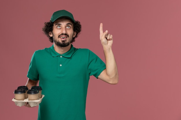 Front view young male courier in green uniform and cape holding brown delivery coffee cups posing and thinking on the pink background service uniform delivery worker job