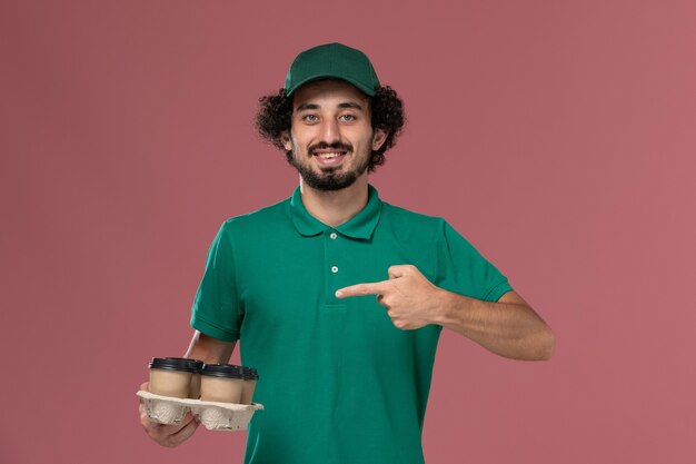 Front view young male courier in green uniform and cape holding brown delivery coffee cups smiling on light-pink background service uniform delivery worker job