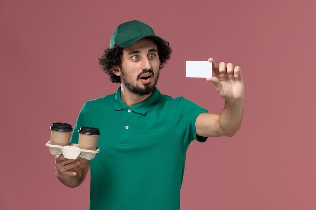 Front view young male courier in green uniform and cape holding delivery coffee cups and card on the pink background service job uniform delivery worker male