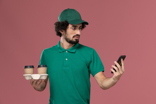 緑の制服を着た若い男性の宅配便とピンクの背景のサービス制服配達労働者に配達コーヒーカップと彼の電話を保持している岬の正面図