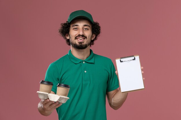 Front view young male courier in green uniform and cape holding delivery coffee cups and notepad on pink background male service job uniform delivery