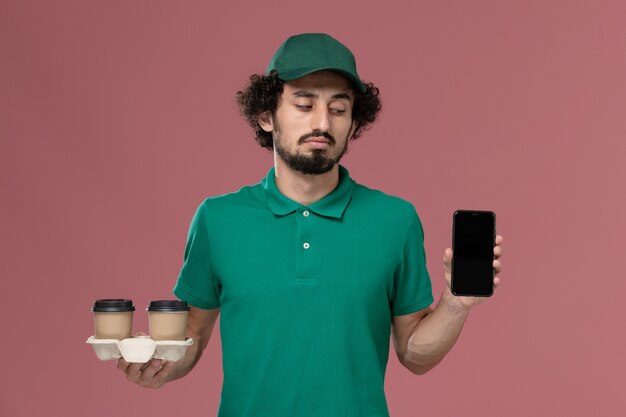 正面図緑の制服を着た若い男性の宅配便と淡いピンクの背景に配達コーヒーカップと電話を保持している岬サービス仕事制服配達