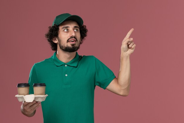 Front view young male courier in green uniform and cape holding delivery coffee cups on pink background service job uniform delivery worker