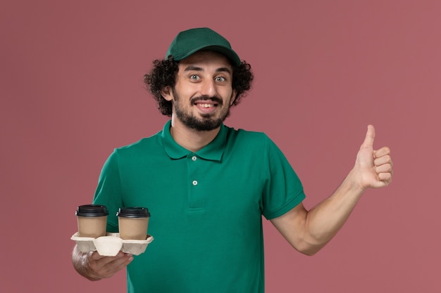 Front view young male courier in green uniform and cape holding delivery coffee cups on the pink background service job uniform delivery