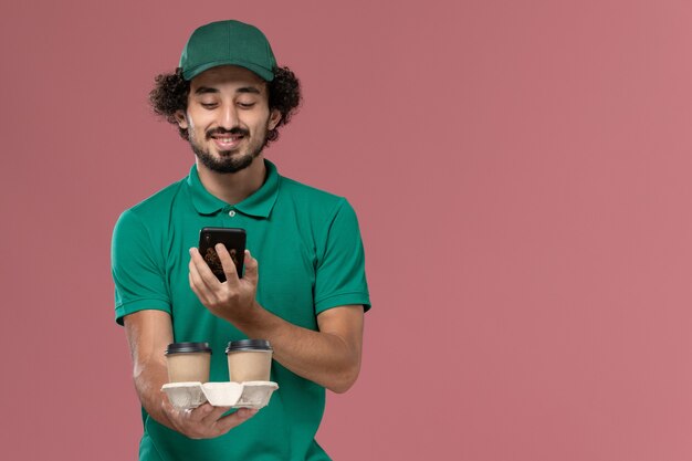 Front view young male courier in green uniform and cape holding delivery coffee cups taking shot of them on the pink background service job uniform delivery worker