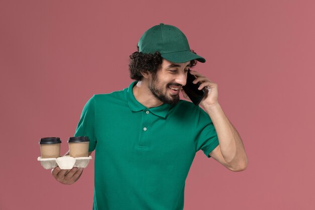Вид спереди молодой мужчина-курьер в зеленой форме и накидке, держащий доставку кофейных чашек, разговаривает по телефону на розовом фоне, служба доставки униформы
