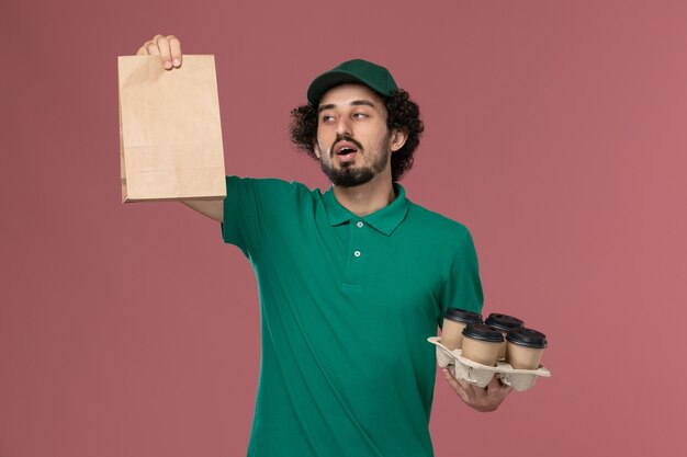 Front view young male courier in green uniform and cape holding delivery coffee cups with food package on light-pink background service uniform delivery job