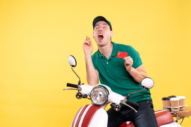 Front view young male courier in green uniform holding bank card on yellow 