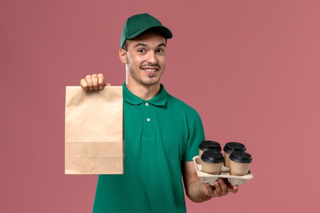 Front view young male courier in green uniform holding brown coffee cups and food package on pink desk