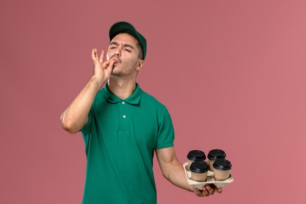 Front view young male courier in green uniform holding brown coffee cups on light-pink background