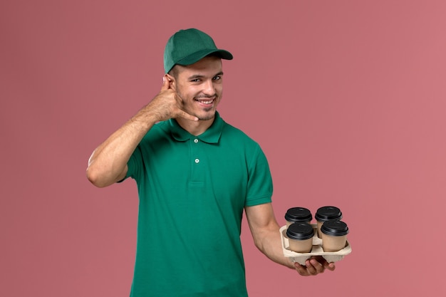 Front view young male courier in green uniform holding brown coffee cups on light pink background