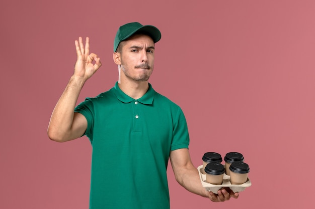 Front view young male courier in green uniform holding brown coffee cups on light pink background 