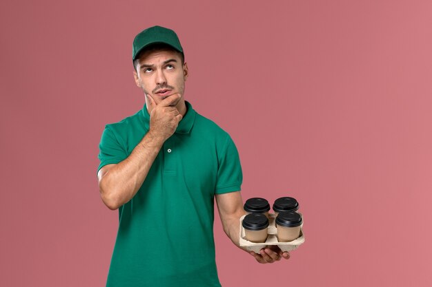 Front view young male courier in green uniform holding brown coffee cups thinking on light pink desk