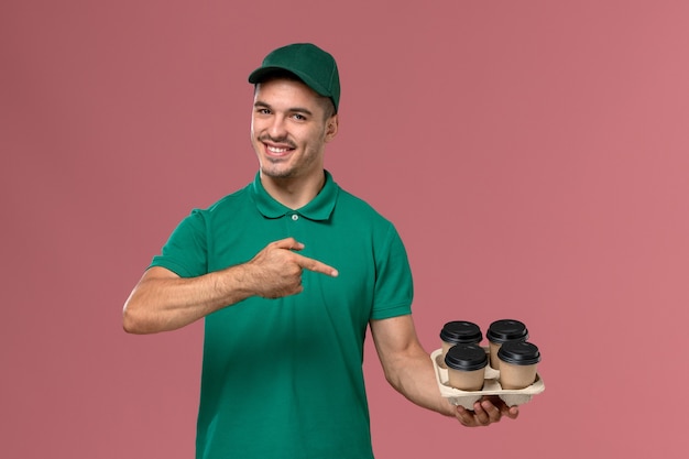 Front view young male courier in green uniform holding brown coffee cups with a smile on pink desk