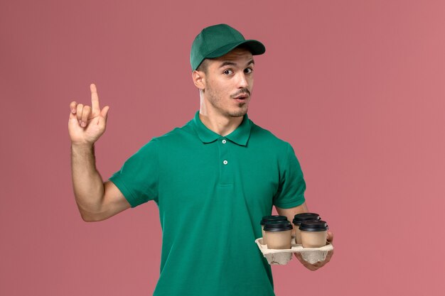 Вид спереди молодой мужчина-курьер в зеленой форме, держащий кофейные чашки, поднимающий палец на розовом фоне