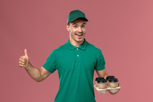 Front view young male courier in green uniform holding coffee cups rejoicing on pink background