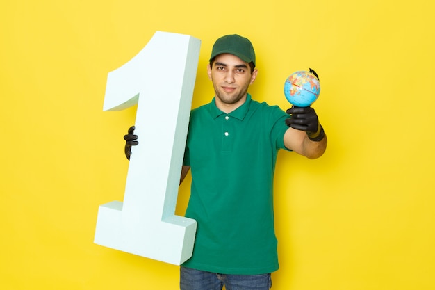 Бесплатное фото Вид спереди молодой курьер-мужчина в зеленой рубашке, зеленая кепка, черные перчатки, держит глобус и номер один на желтом