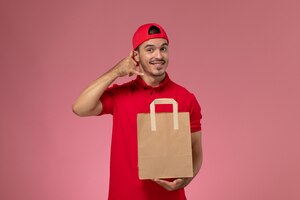 front view young male courier in red uniform cape holding paper food package on the pink background.