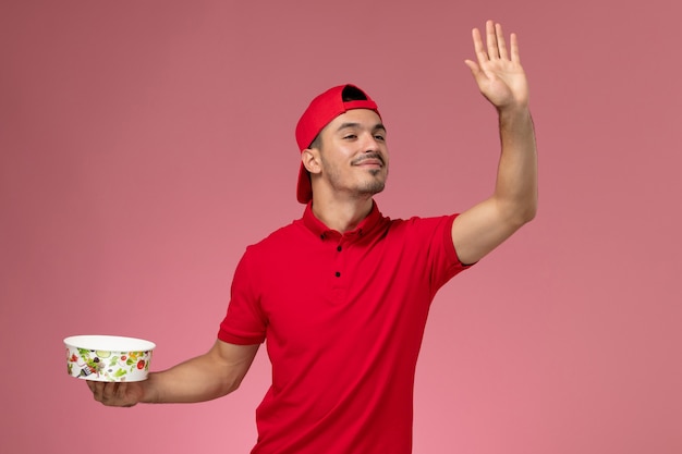 Front view young male courier in red uniform cape holding delivery bowl and waving on light-pink background.