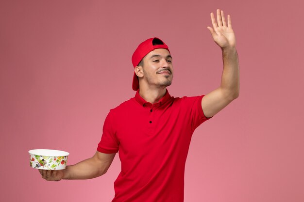 Front view young male courier in red uniform cape holding delivery bowl and waving on light-pink background.