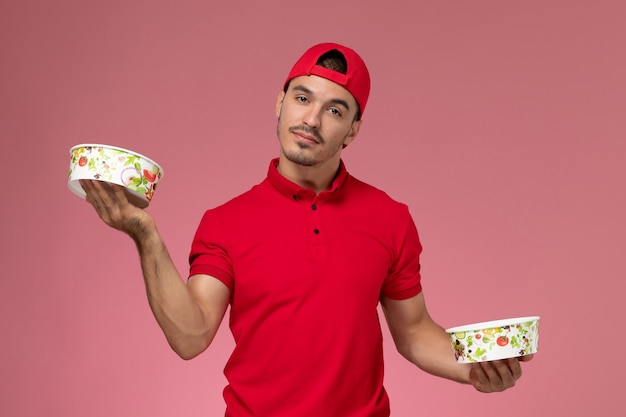 Front view young male courier in red uniform cape holding delivery bowls on light pink background.