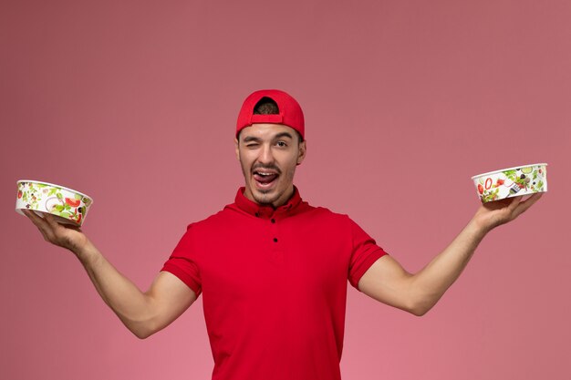 Front view young male courier in red uniform cape holding delivery bowls winking on light pink background.