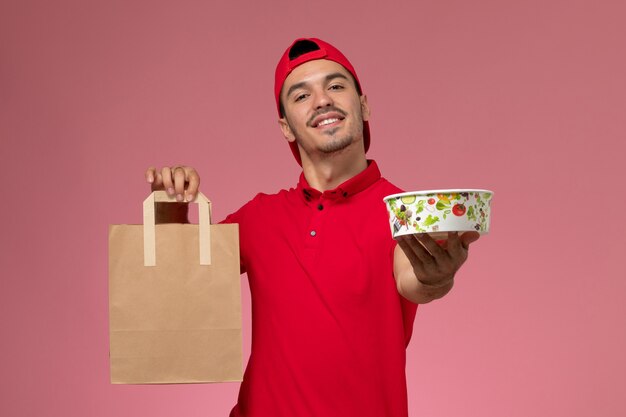 Front view young male courier in red uniform cape holding food package and bowl on pink background.