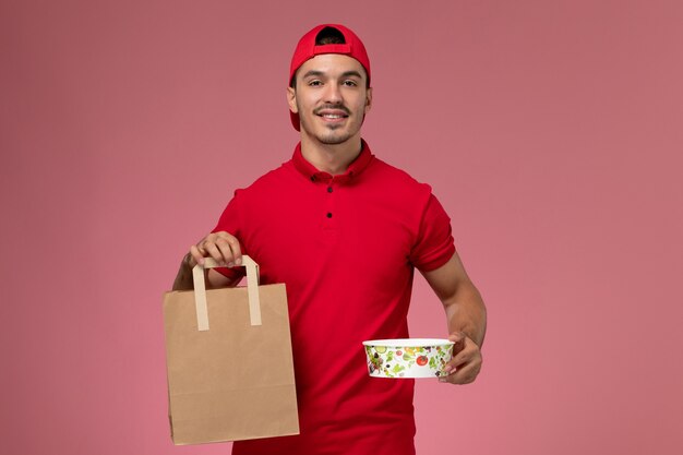Вид спереди молодой мужской курьер в красной форме накидки, держащей пакет и миску с улыбкой на розовом фоне.