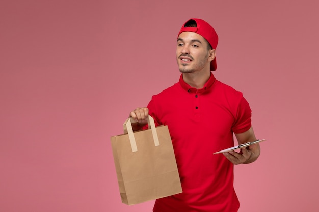 淡いピンクの背景に食品パッケージとメモ帳を保持している赤い制服の岬の正面図の若い男性の宅配便。