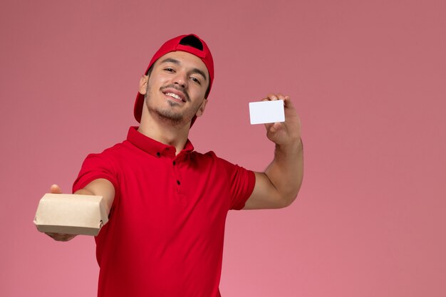 분홍색 배경에 흰색 카드와 함께 작은 배달 패키지를 들고 빨간 유니폼 케이프에서 전면보기 젊은 남성 택배.