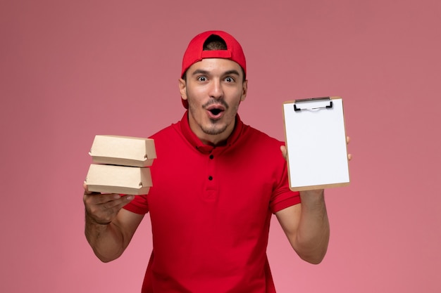 Front view young male courier in red uniform cape holding little packages with food and notepad on light-pink background.