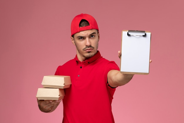 Front view young male courier in red uniform cape holding little packages with food and notepad on light-pink background.