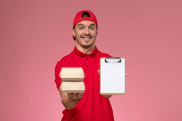 Front view young male courier in red uniform cape holding notepad and little package with food on pink desk.