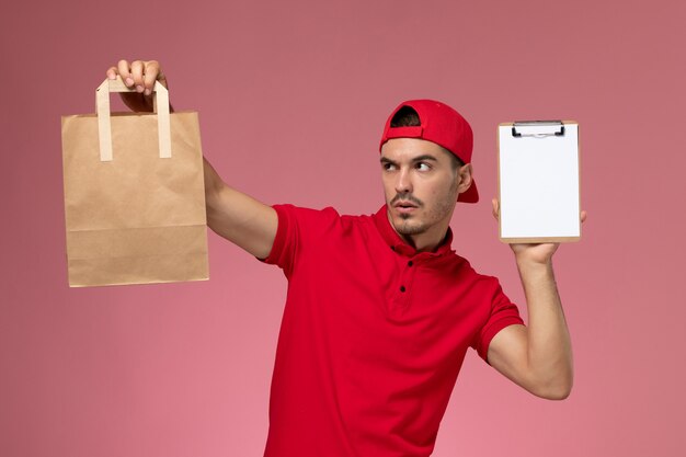 ピンクの背景に食品パッケージとメモ帳を保持している赤い制服ケープの正面図若い男性宅配便。
