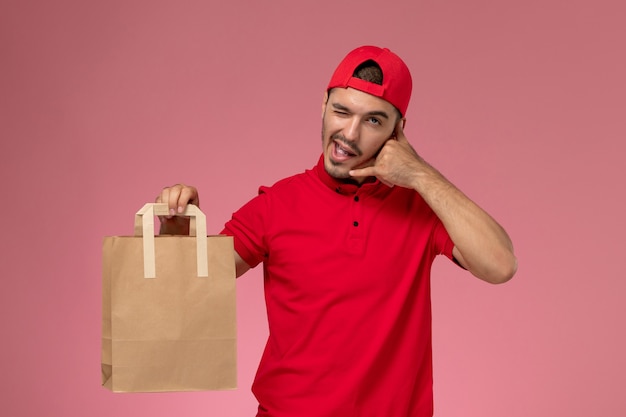 Front view young male courier in red uniform cape holding paper food package on the light pink background.
