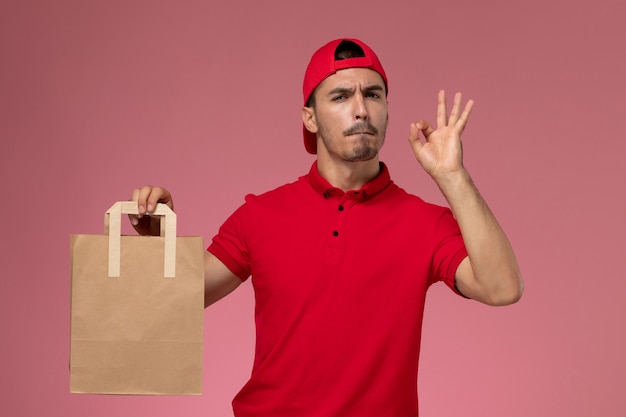 Front view young male courier in red uniform cape holding paper food package on pink background.