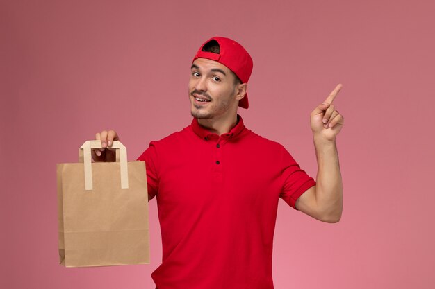 ピンクの背景に紙の食品パッケージを保持している赤い制服の岬の正面図若い男性宅配便。