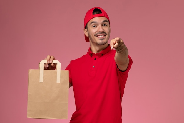 Front view young male courier in red uniform cape holding paper food package and smiling on pink background.