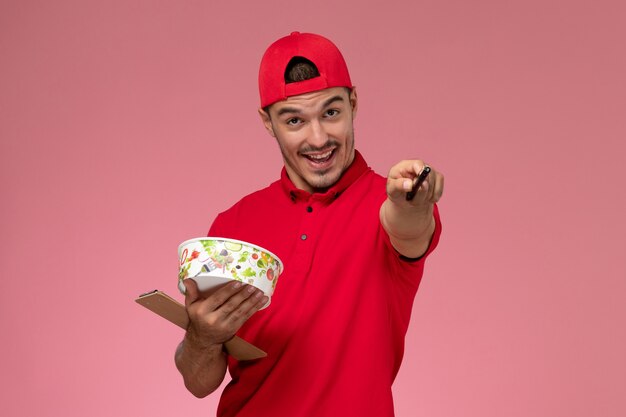 Front view young male courier in red uniform cape holding round delivery bowl and notepad on pink background.